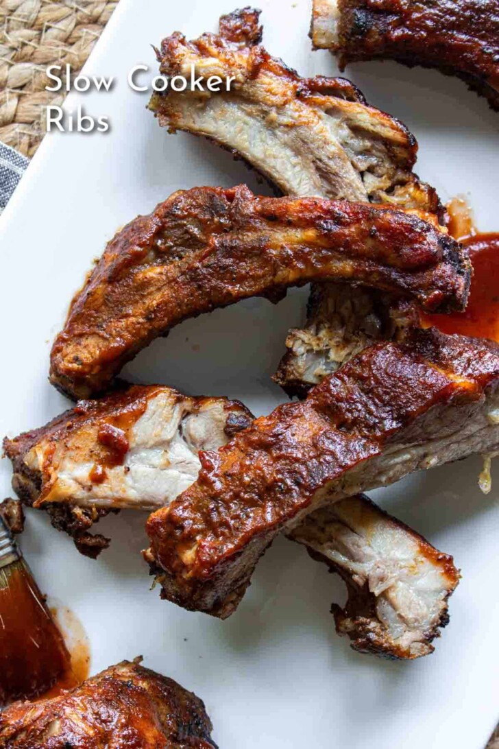 Close-up of several pieces of Slow Cooker Ribs coated in barbecue sauce on a white plate. The text